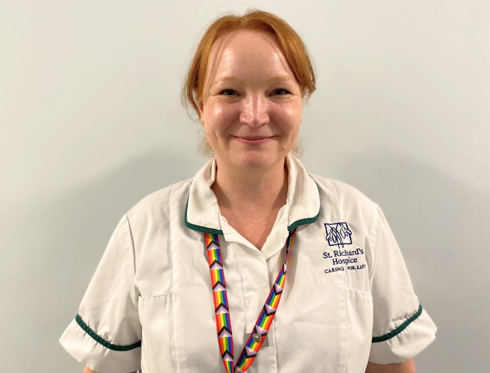Sally pictured standing in front of a plain white wall at the hospice. She is wearing a white Occupational Therapist's uniform, with dark green edging. She also has a lanyard on in the colours of the progress Pride flag. She is smiling.