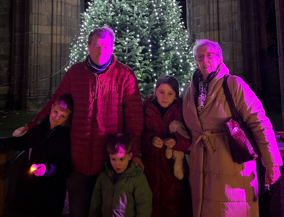 The Bader family stand together in front of the St Richard's Hospice Lights of Love Christmas tree, which is lit with lots of silvery white fairy lights. It is dark outside, and the family are illuminated in purply pink light.
