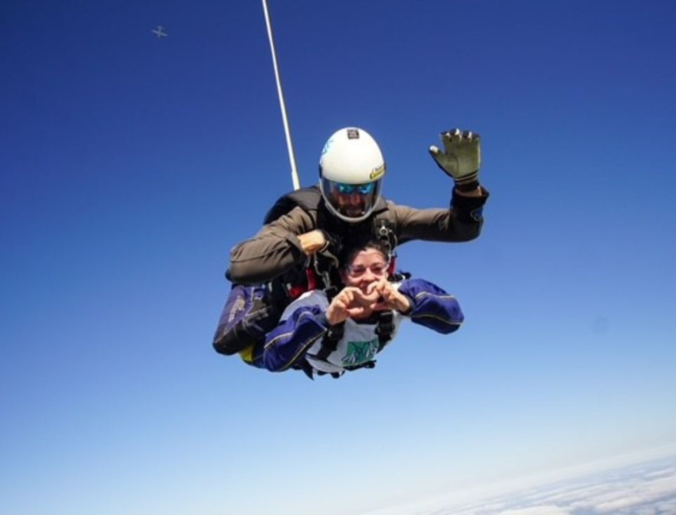 A woman taking part in a skydive, free falling with a parachute expert above her surrounded by blue sky.