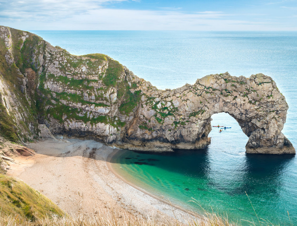 A sandy beach with blue water and a rock face that has been eroded into an arch.