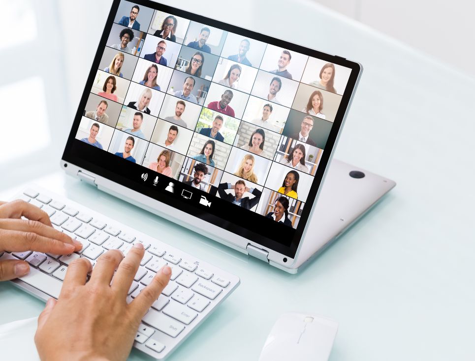 An image of a video call taking place on a laptop with lots of people on the call. The laptop is on a white table.
