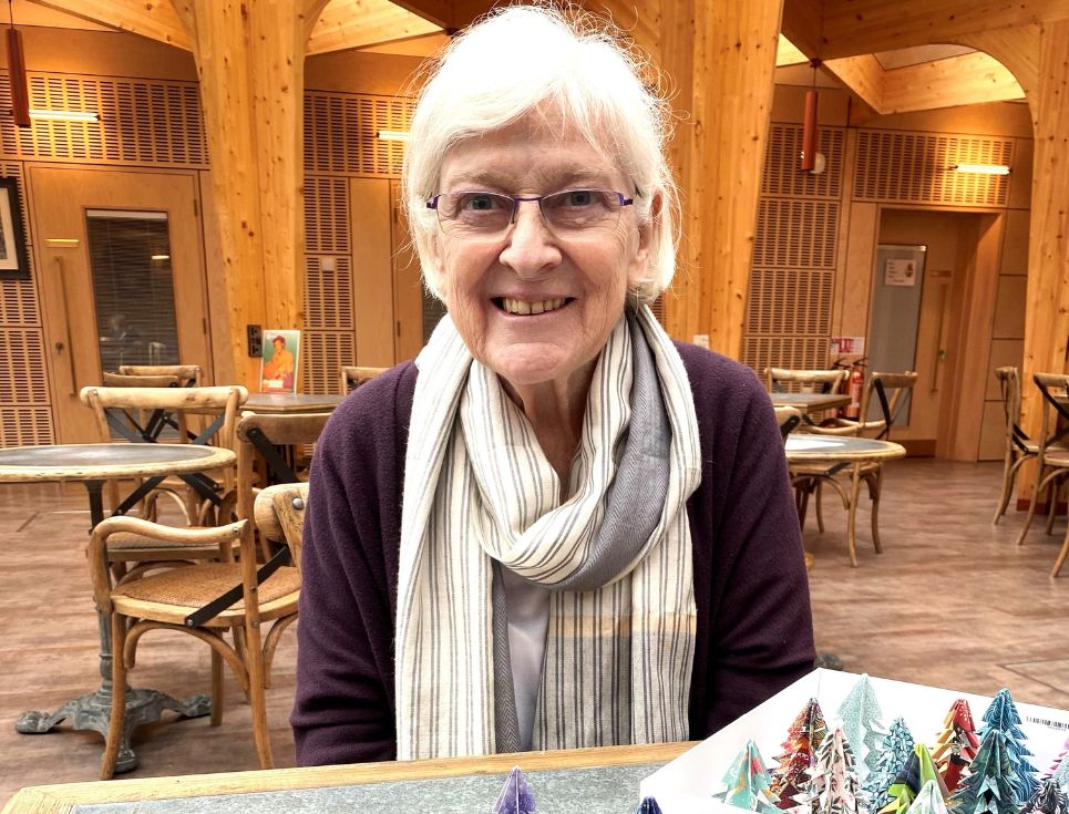 Vanessa pictured at the hospice with a collection of colourful paper Christmas trees. Vanessa is wearing a dark purple jumper and a cream and grey striped scarf. She is smiling.