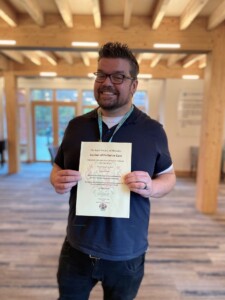 Andy stands in the hospice's Reception area holding up a certificate. He is smiling.