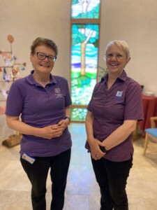 Chaplains Clare and Sarah stand side-by-side in the Sacred Space at St Richard's Hospice. Behind them is a tall, slim, stained glass window featuring a tree. They are both wearing purple tops featuring the hospice logo, and black trousers. They are both smiling.