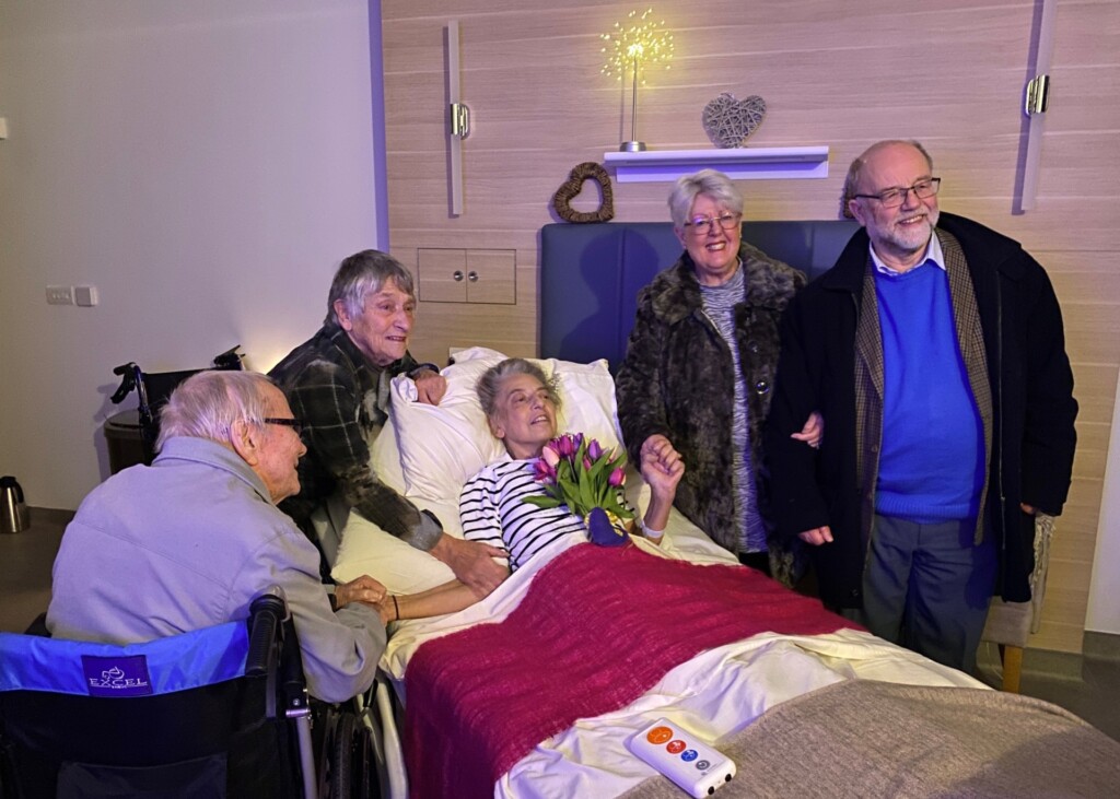 Sarah sits in her bed at the hospice with a bunch of colourful tulips resting on her chest. She is surrounded by her parents and her husband's parents, who are all smiling.