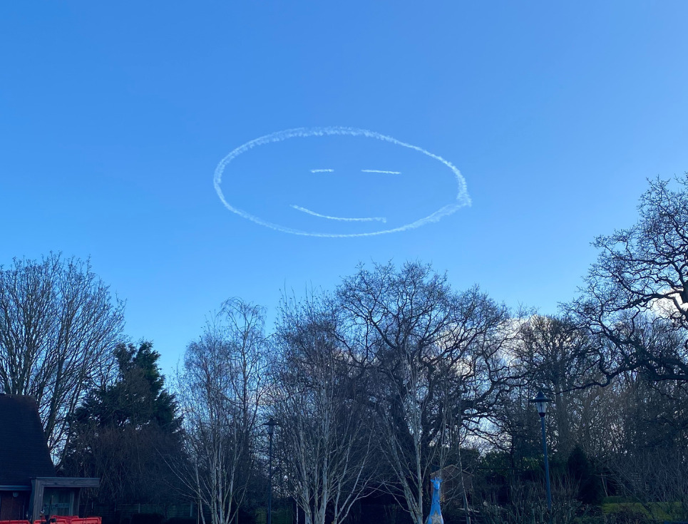 A smiley face drawn in the sky using a plan's vapour trails. The sky is bright blue and trees line the bottom half of the image, taken in the hospice's gardens.