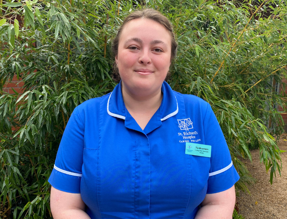 Tia, pictured standing in front of a green shrub in the hospice's courtyard garden. She is wearing a bright blue nurses' uniform and is smiling.