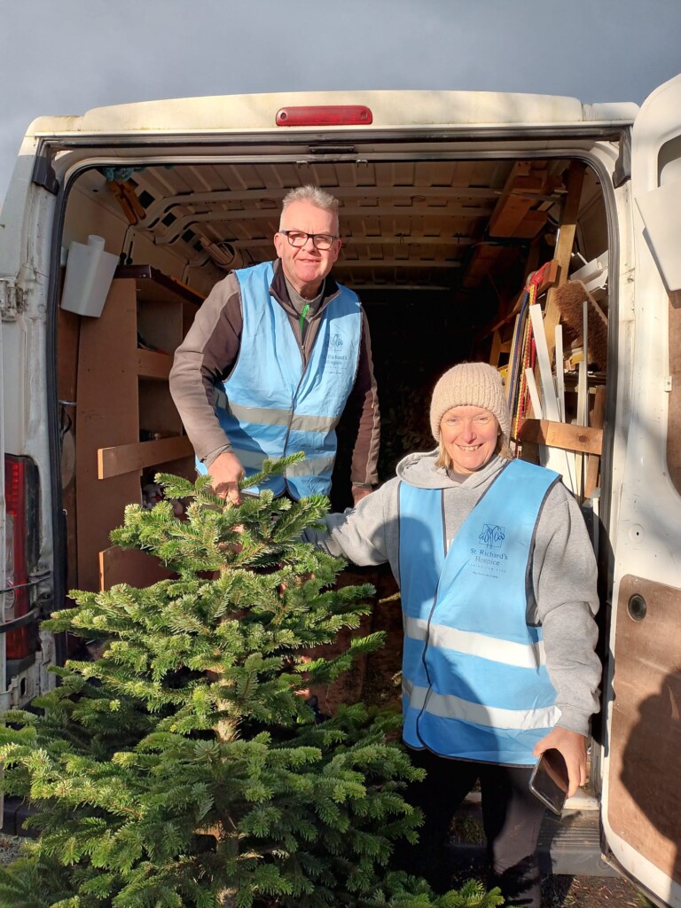 Two people stand at the back of a van holding a Christmas tree ready to load it into the vehicle. They are both wearing blue high-vis jackets and warm clothes.