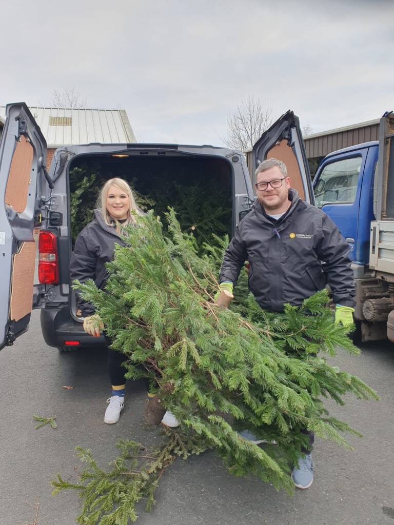 Two volunteers stand behind a van with its doors open. Inside the van are Christmas trees ready for recycling, and the two people stand holding Christmas trees ready to put them into the van.