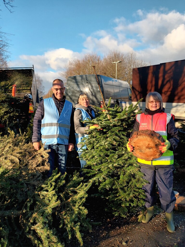 Three people wearing high-vis jackets stand outside with large green Christmas trees. They are part of the hospice's Christmas Tree Recycling Campaign.