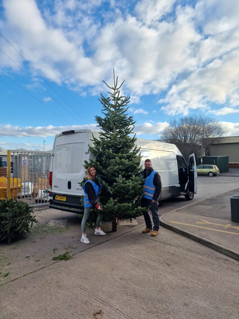 Two people in high-vis jackets stand either side of a giant Christmas tree. Behind them is a large white van and the sky is blue with white clouds.