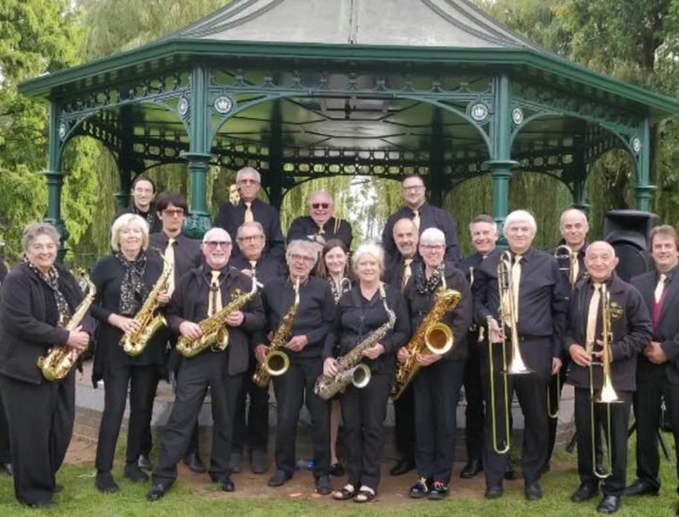 A brass band standing at a bandstand wearing black, holding their instruments.