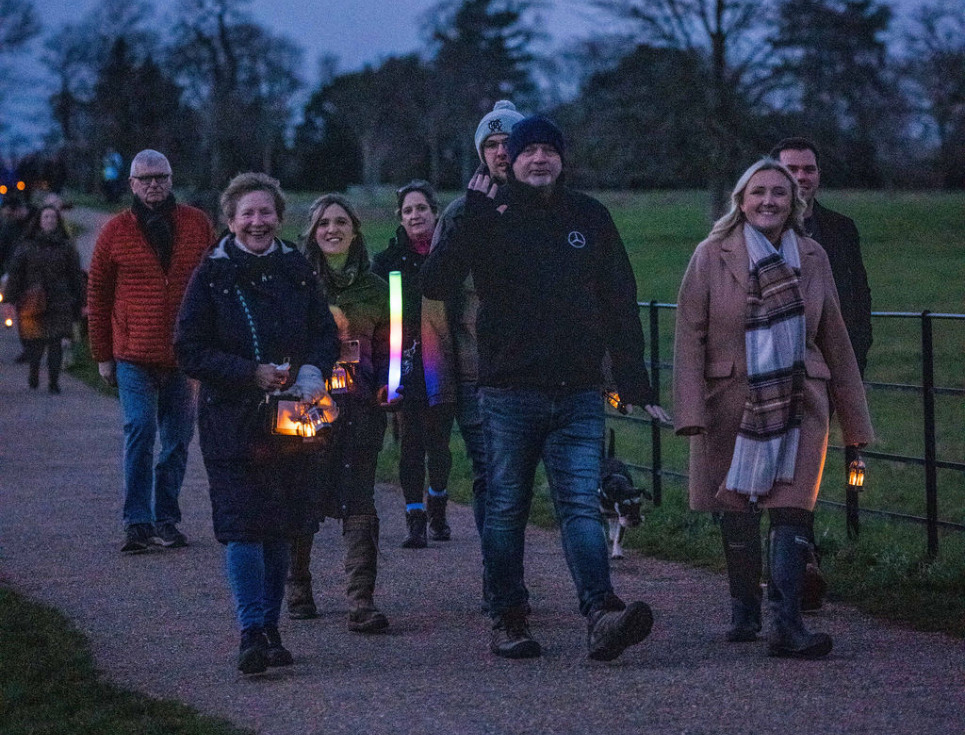 A group of people walk through parkland of Croome Court at twilight. They are holding glowing lanterns and smiling.