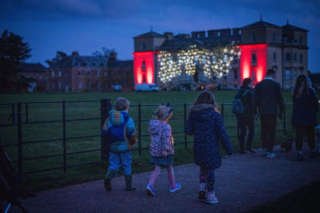 Three children take part in the hospice's Lantern Walk. behind them is the house at Croome illuminated with red and gold lights. It is twilight.