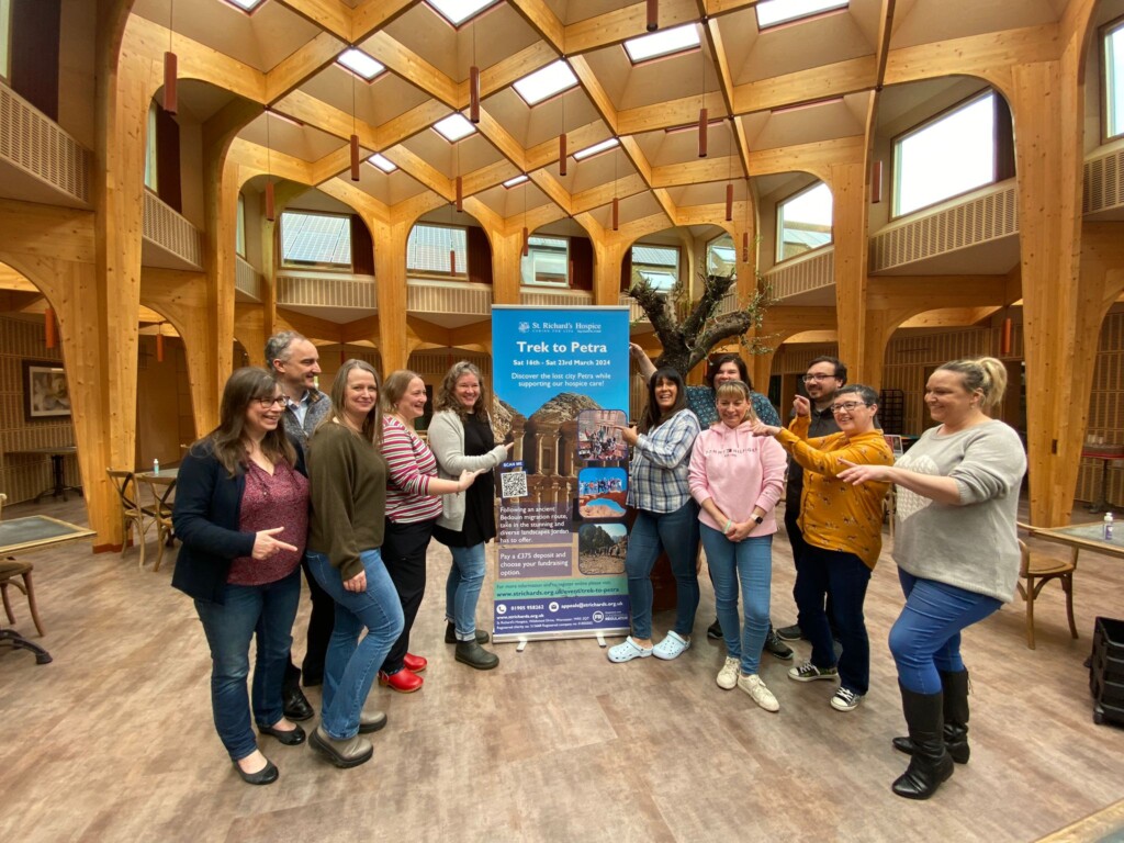 A group of people stand in The Green social space at St Richard's Hospice pointing at a tall banner featuring the Trek to Petra challenge wording and images. 