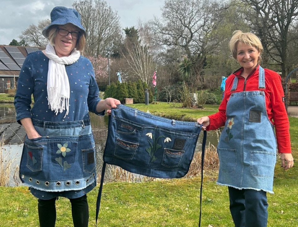 Two people stand together in the hospice gardens holding up a half-apron made from denim between them. They are each wearing handmade denim aprons and smiling.