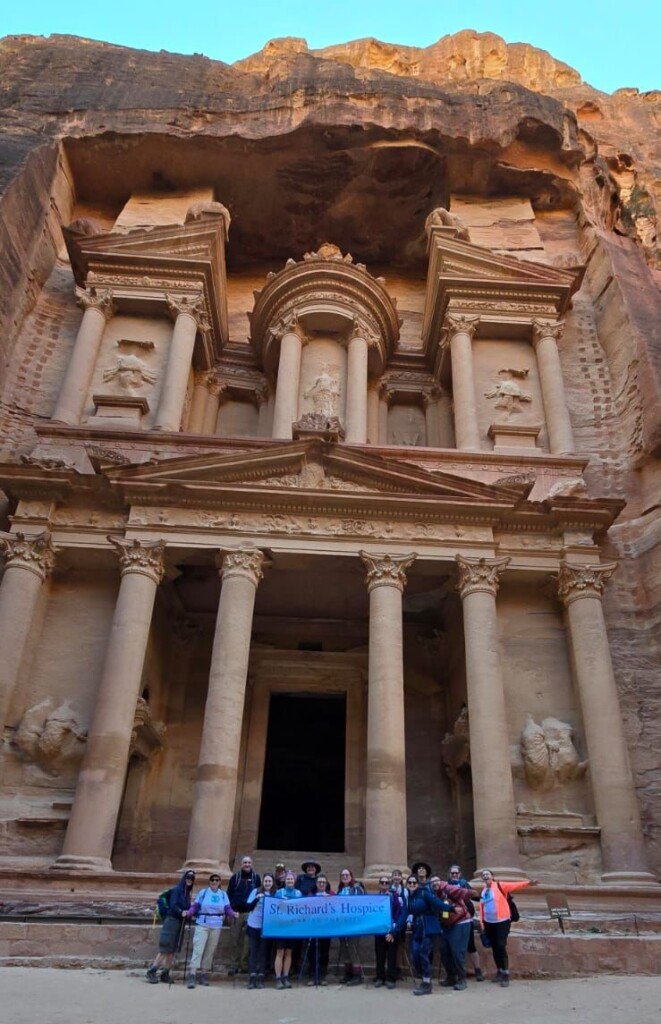 The group of St Richard's trekkers reach the historic city of Petra. The group stand in front of one of the ancient buildings carved into the rock face. The building is very tall, carved into the red coloured stone. 