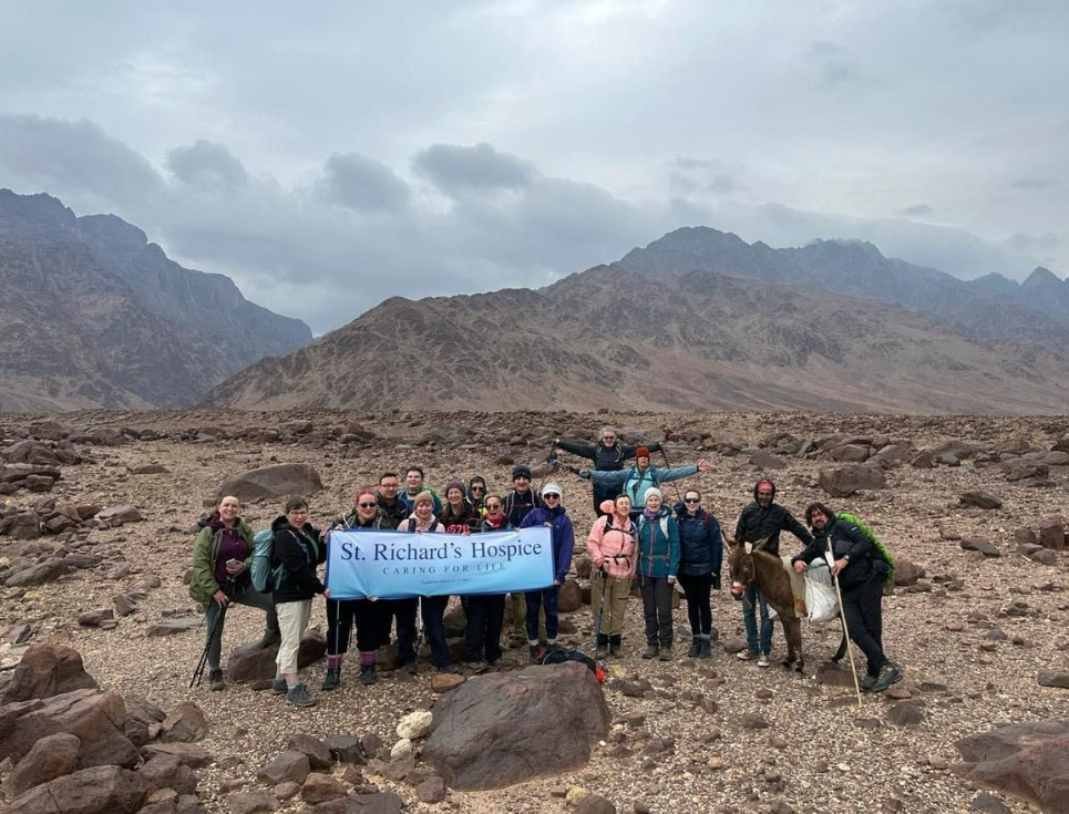 A group of walkers pose for a photo in the Jordanian desert, holding up a banner featuring the St Richard's Hospice logo. Behind them is a spectacular mountain range, and a grey cloudy sky.