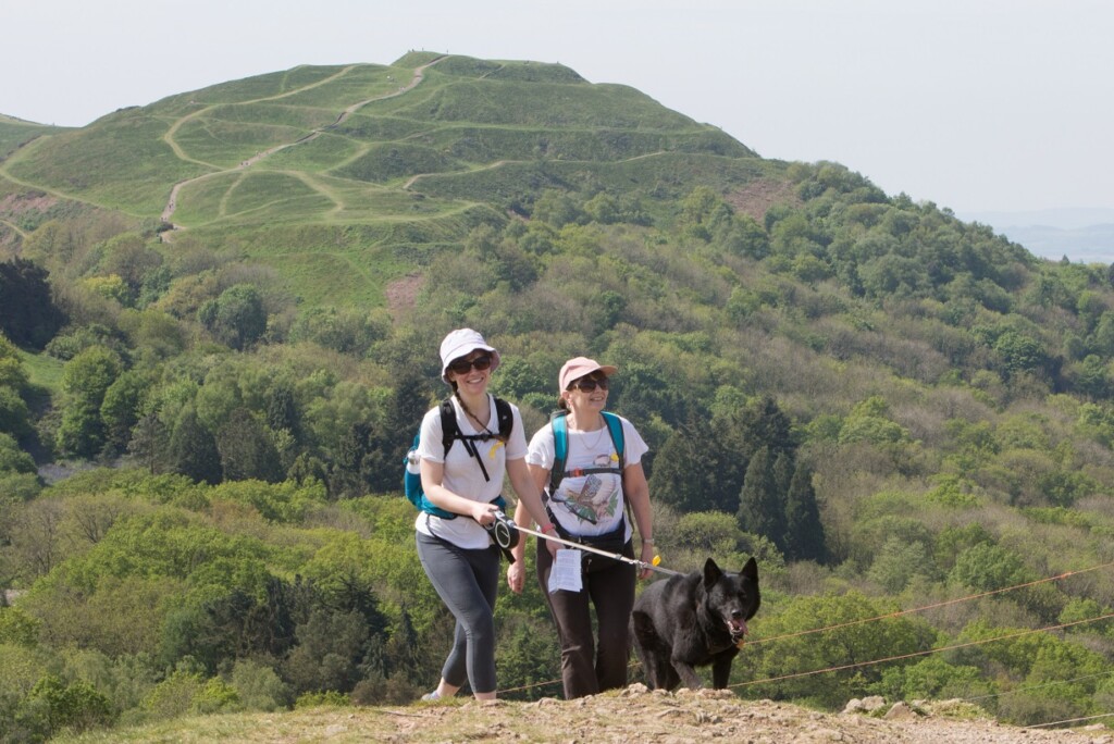 Two people walk over the Malvern Hills during the St Richard's Malvern Hills Walk. The sun is shining and one of the hills is behind them. One of the people is holding a black dog on a lead.