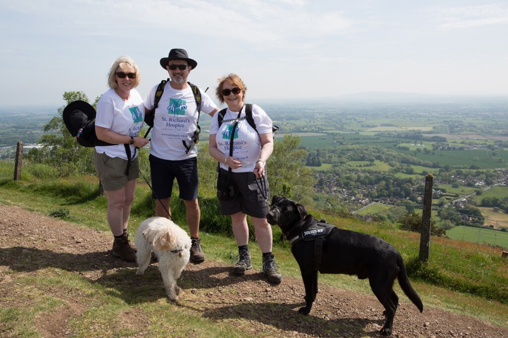 Three people in St Richard's branded t-shirts and shorts stand on the Malvern Hills during the hospice's fundraising walk. Two dogs are in the photo with them.