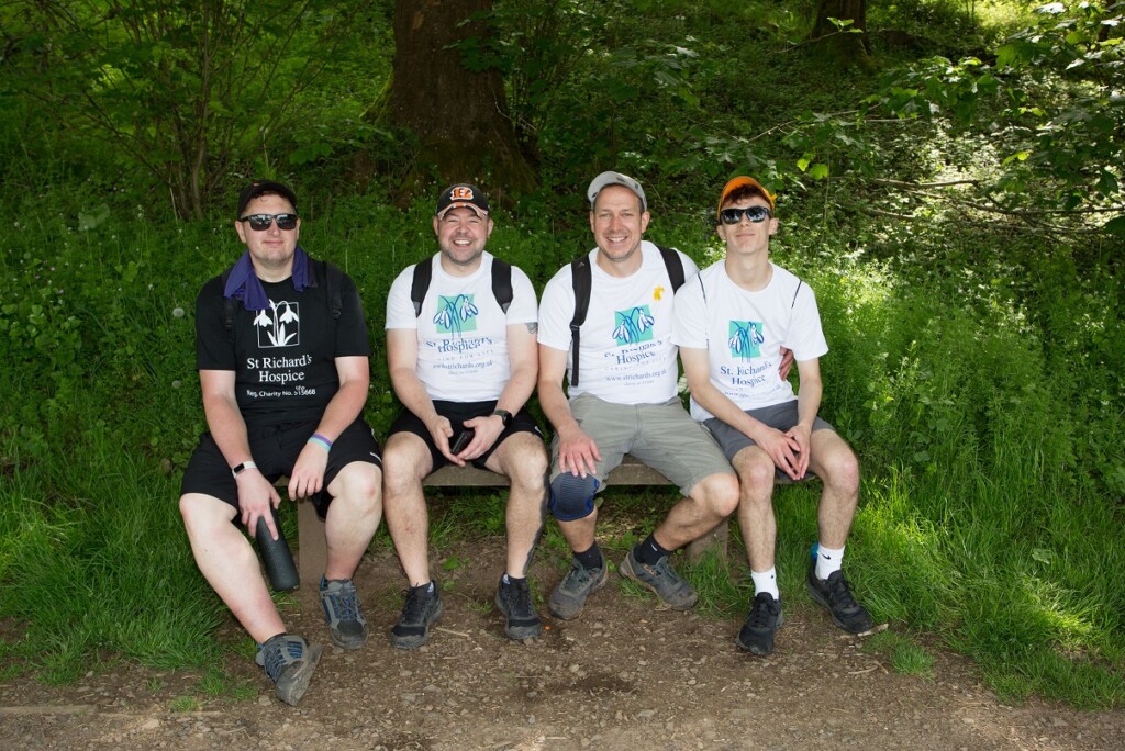 Four people wearing St Richard's branded t-shirts and shorts sit together on a bench during the Malvern Hills Walk. Behind them is green grass and trees. 