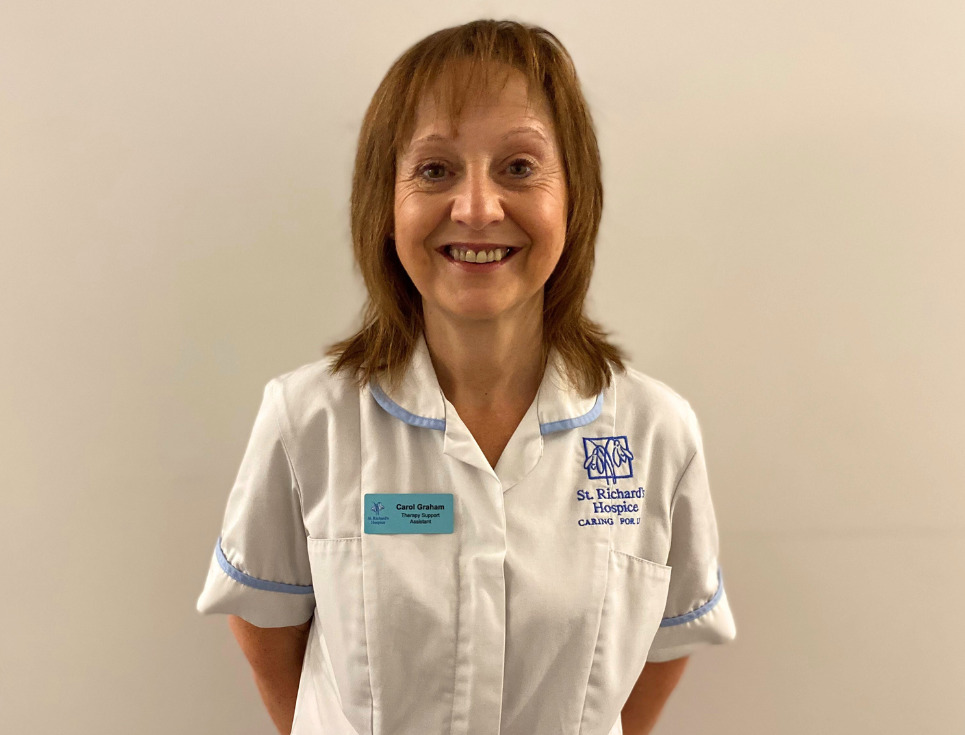 Carol stands in front of a plain white wall and is smiling. She is wearing a white therapy support assistant's uniform.