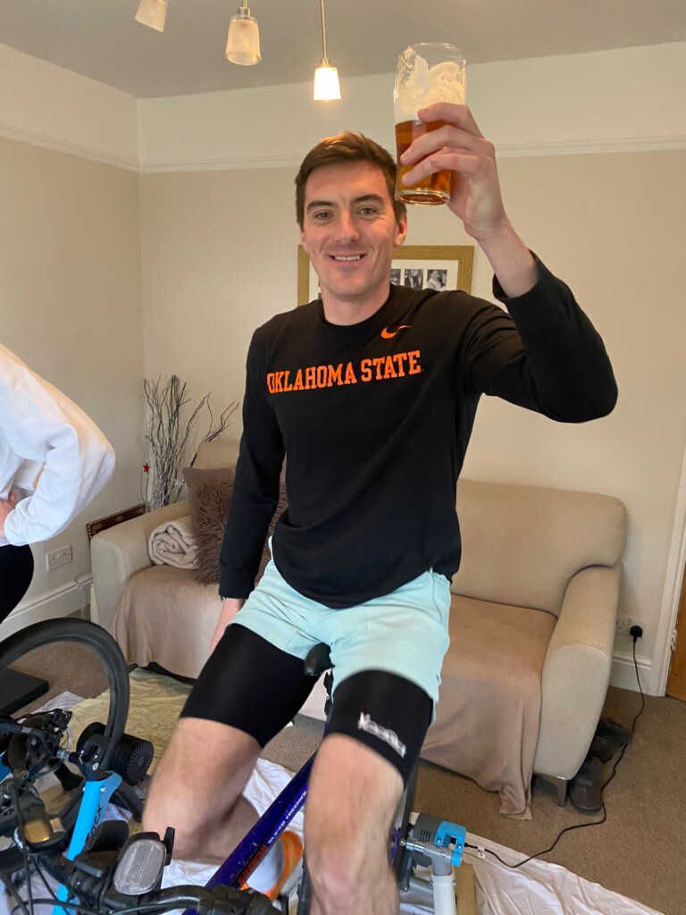 A man raises a pint while cycling on a static bike inside a house.