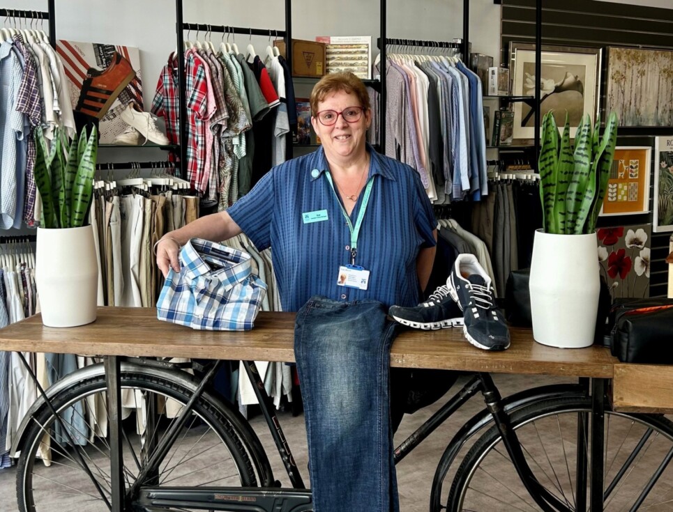 Sue stands in the hospice's store behind a display stand with bicycle wheels underneath holding up a folded shirt. On the display are two potted plants, a pair of jeans, and a pair of trainers.