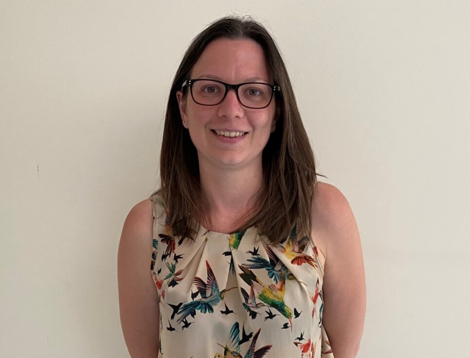 A photo of Rosie, who has shoulder-length dark brown hair, stands in front of a white wall. She is smiling and wearing a short-sleeved top with a colourful bird print and black trousers.