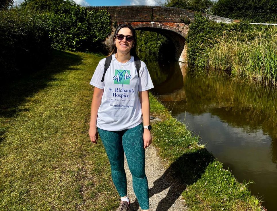 Sofia, dressed in blue green leggings and a white St Richard's branded t-shirt, stands on a canal towpath next to a canal. Behind her is a picturesque bridge and blue sky.