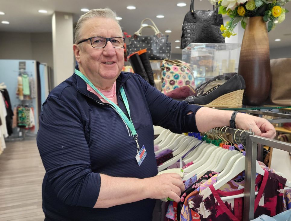 Volunteer Dexter arranging a rail of clothing in a hospice shop. He is smiling.