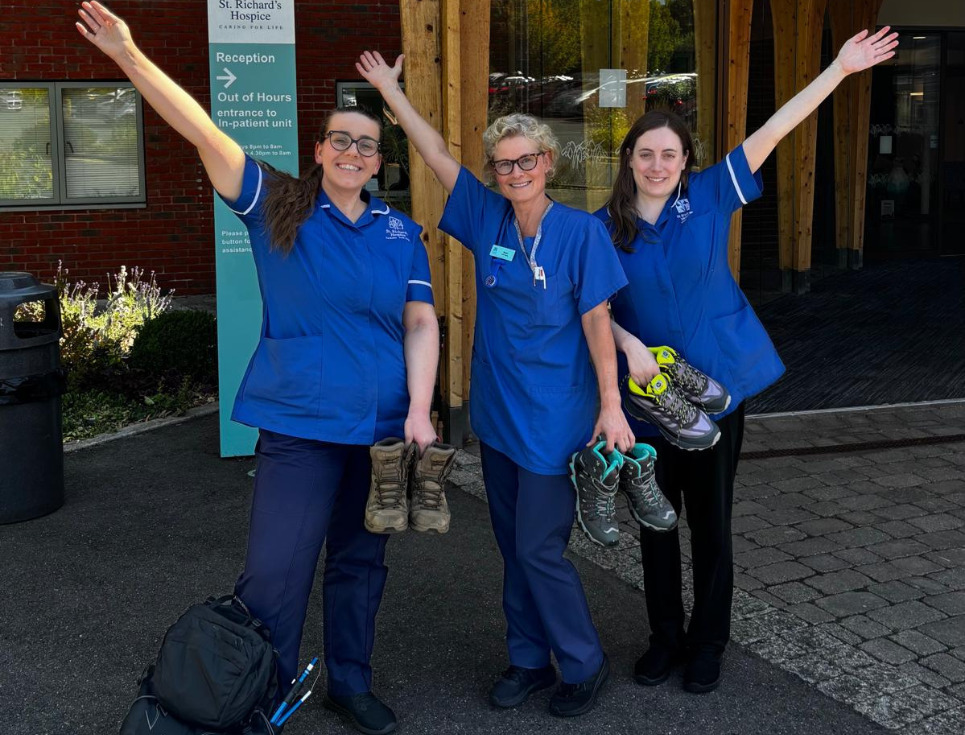 Three hospice nurses wearing blue uniforms stand outside the hospice. They are holding their walking boots and are waving their free arm.